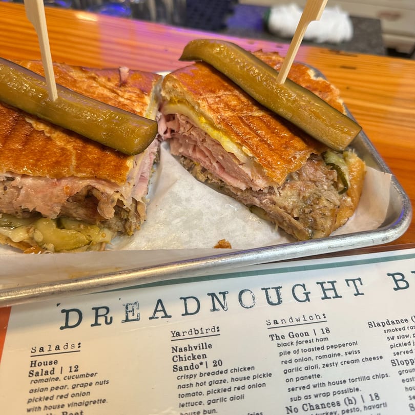 Two halves of a Cuban sandwich with pickles on top, served on a metal tray with a menu.